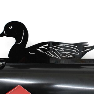 May include: Black metal mailbox topper shaped like a duck in a swimming position. The duck is facing the right side of the image. The mailbox topper is designed to be placed on top of a standard mailbox.