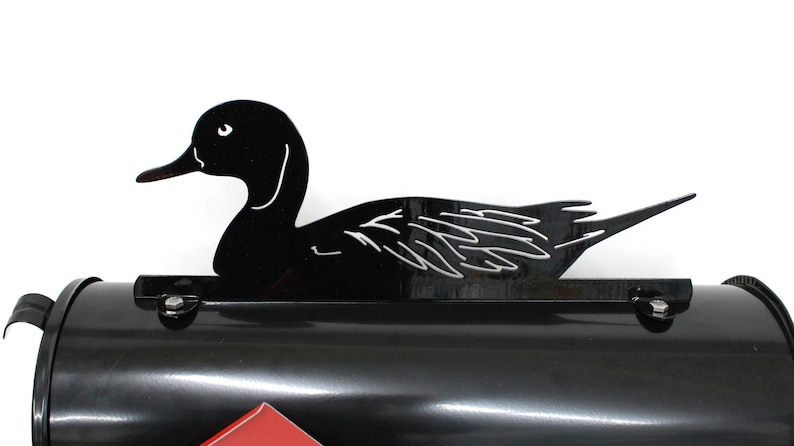 May include: Black metal mailbox topper shaped like a duck in a swimming position. The duck is facing the right side of the image. The mailbox topper is designed to be placed on top of a standard mailbox.