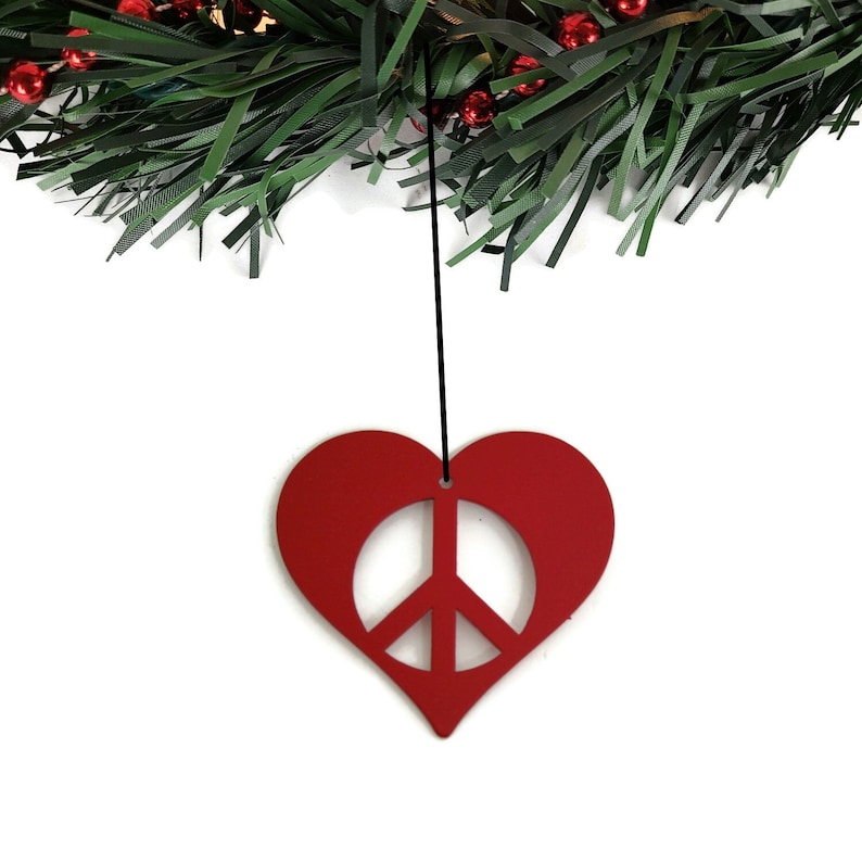 May include: A red heart-shaped ornament with a peace sign cut out in the center, hanging from a green Christmas wreath.