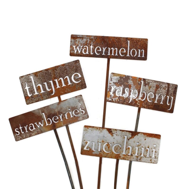 May include: A collection of weathered metal garden markers with the names of various plants in white lettering. The markers include "thyme", "strawberries", "watermelon", "raspberry", and "zucchini". The metal has a rusty, aged appearance.