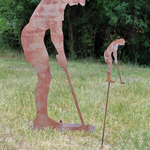 May include: Two rusty metal golf figures, one large and one small, are positioned on a grassy lawn. The larger figure is bent over, holding a golf club. The smaller figure is on a stake, also holding a club. Green trees are in the background.