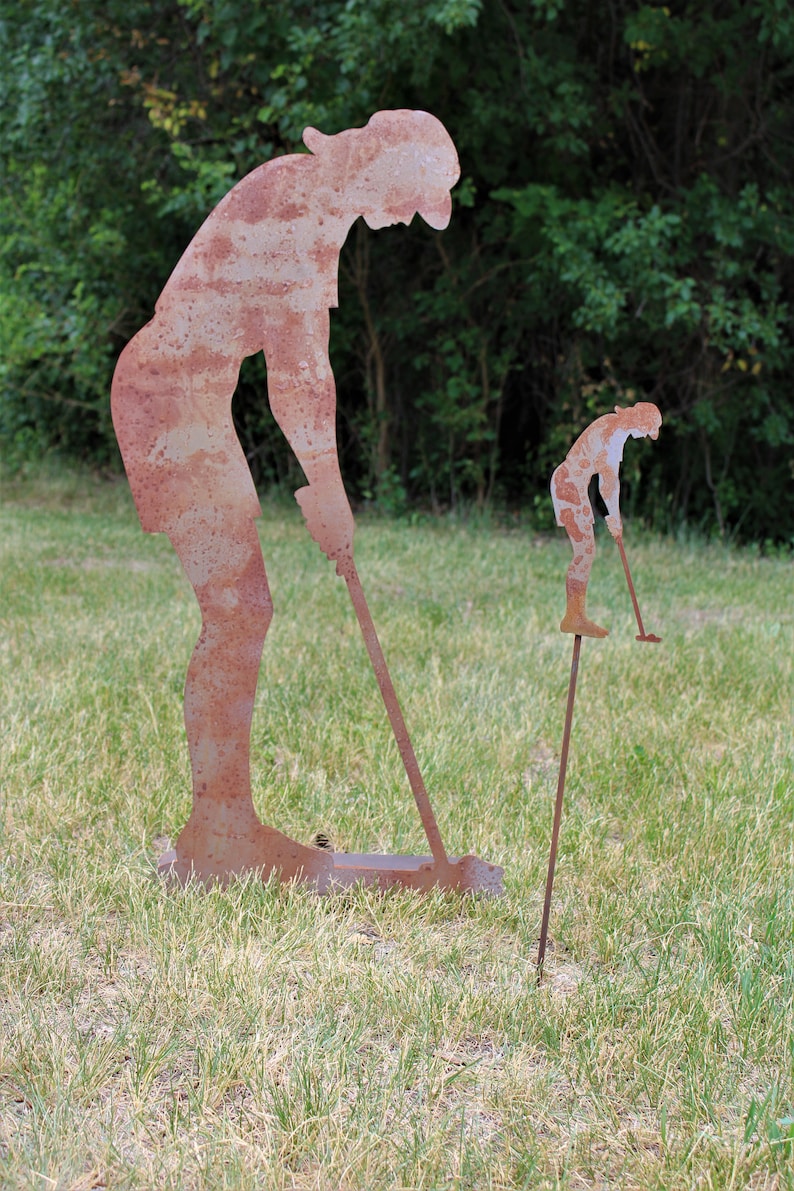 May include: Two rusty metal golf figures, one large and one small, are positioned on a grassy lawn. The larger figure is bent over, holding a golf club. The smaller figure is on a stake, also holding a club. Green trees are in the background.