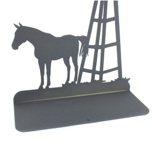 Op de afbeelding: Een zwart metalen silhouet van een paard dat naast een windmolen staat met een plank eronder. Het silhouet is aan een muur gemonteerd.