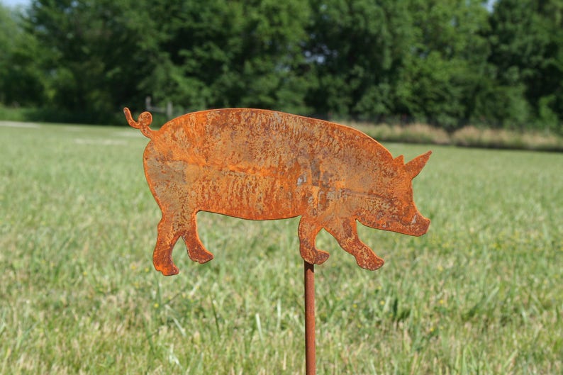 May include: A rusty metal pig silhouette garden stake. The pig is facing right and has a curled tail. The stake is inserted into the ground.