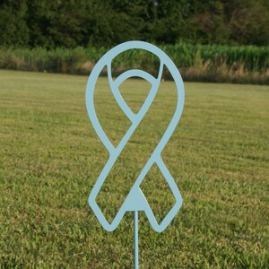 May include: A light blue metal ribbon yard stake. The ribbon is a cutout design with a pointed bottom and a stake for placement in the ground. The background is a grassy field with trees.