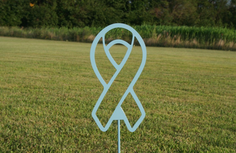 May include: A light blue metal ribbon yard stake. The ribbon is a cutout design with a pointed bottom and a stake for placement in the ground. The background is a grassy field with trees.