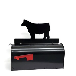 May include: Black metal mailbox with a red flag and a decorative black cow silhouette. The mailbox has a curved top and a rectangular shape, suitable for rural or farm-themed decor.