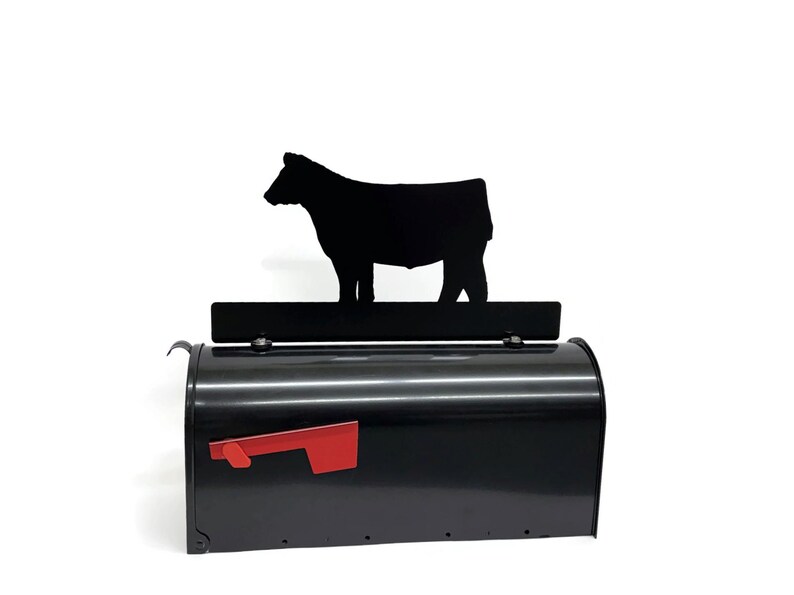 May include: Black metal mailbox with a red flag and a decorative black cow silhouette. The mailbox has a curved top and a rectangular shape, suitable for rural or farm-themed decor.
