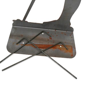 May include: A weathered metal garden stake featuring a silhouette of a person with a bag, along with three metal stakes. The metal has a rusty appearance, suggesting outdoor use. The design is simple and functional.