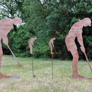 May include: Four rusty metal golf player silhouettes are positioned on a grassy lawn. The figures are in various stages of a golf swing, with clubs extended. Green trees form the background, creating a natural setting for the outdoor art.