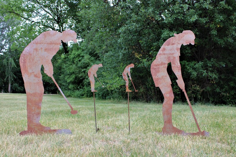 May include: Four rusty metal golf player silhouettes are positioned on a grassy lawn. The figures are in various stages of a golf swing, with clubs extended. Green trees form the background, creating a natural setting for the outdoor art.