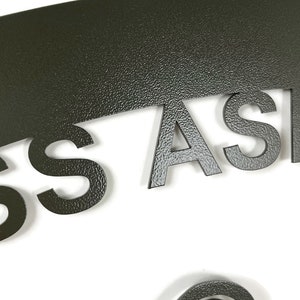 May include: A dark green metal sign with the word "ASSA" cut out in the shape of a half circle. The sign has a textured surface.