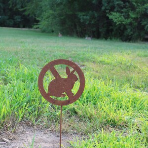May include: A rusty metal sign with a red circle and a diagonal line through it. The sign has a silhouette of a rabbit inside the circle. The sign is on a metal stake and is stuck in the ground.