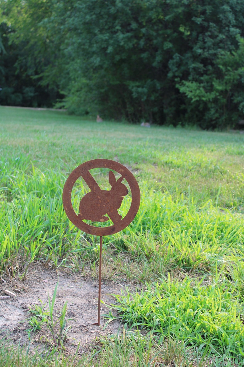 May include: A rusty metal sign with a red circle and a diagonal line through it. The sign has a silhouette of a rabbit inside the circle. The sign is on a metal stake and is stuck in the ground.