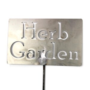 May include: A rectangular, silver-colored metal garden marker with the words "Herb Garden" cut out. The marker is attached to a thin, dark metal stake, perfect for labeling herbs in a garden.