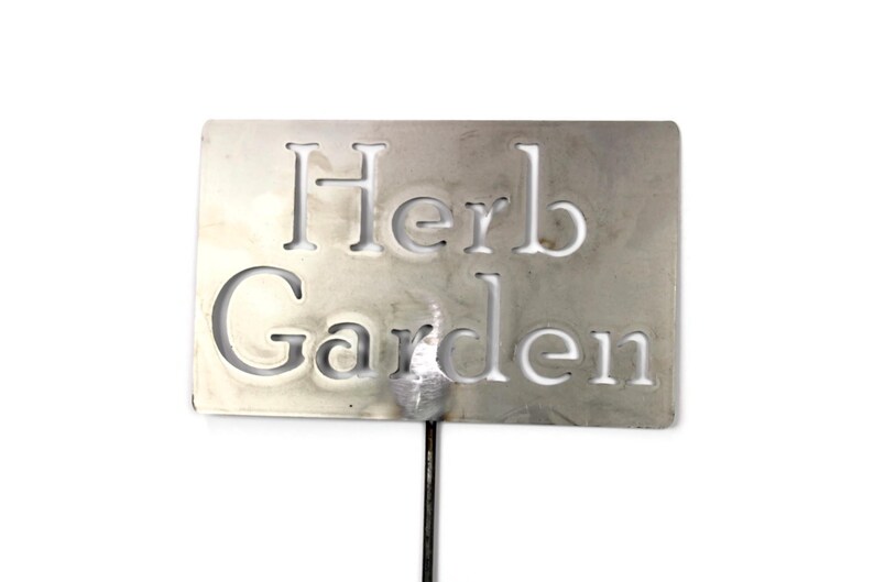 May include: A rectangular, silver-colored metal garden marker with the words "Herb Garden" cut out. The marker is attached to a thin, dark metal stake, perfect for labeling herbs in a garden.