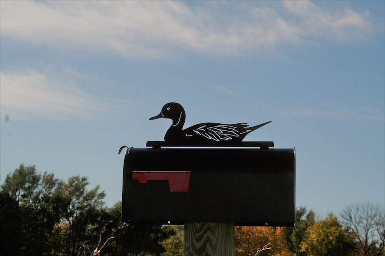 May include: Black metal mailbox with a duck silhouette on top. The mailbox is mounted on a wooden post.