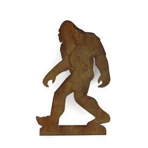 May include: A rusty brown metal silhouette of Bigfoot walking. The figure has a large, upright posture with a defined head, arms, and legs. The metal has a weathered appearance, suggesting outdoor use or an aged aesthetic.