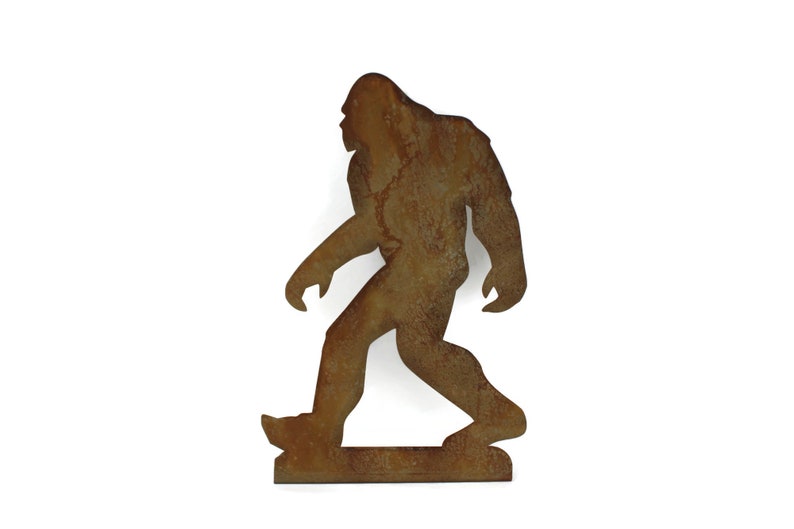 May include: A rusty brown metal silhouette of Bigfoot walking. The figure has a large, upright posture with a defined head, arms, and legs. The metal has a weathered appearance, suggesting outdoor use or an aged aesthetic.