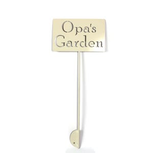 May include: A beige metal garden stake with the words "Opa's Garden" cut out. The sign is rectangular and attached to a thin pole with a rounded base. The background is white.