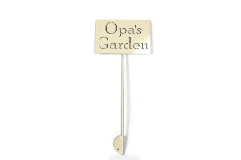 May include: A beige metal garden stake with the words "Opa's Garden" cut out. The sign is rectangular and attached to a thin pole with a rounded base. The background is white.