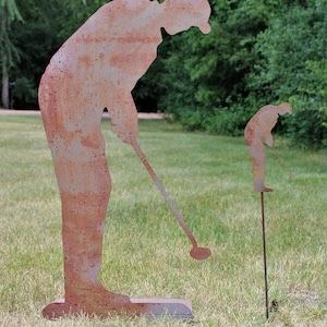 May include: Two rusty metal sculptures of golfers in a grassy outdoor setting. One is a full-sized figure, and the other is a smaller version on a stake. The sculptures depict a golfer in the act of swinging a golf club.