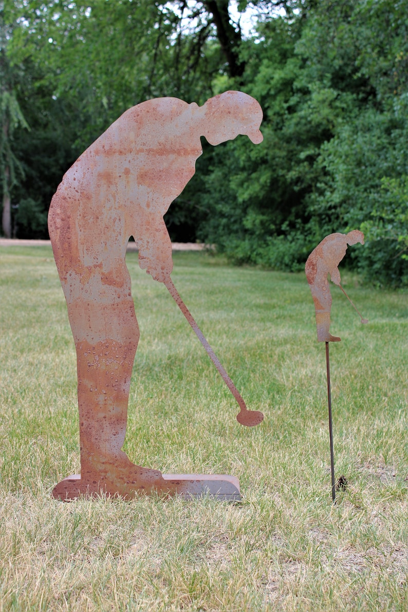 May include: Two rusty metal sculptures of golfers in a grassy outdoor setting. One is a full-sized figure, and the other is a smaller version on a stake. The sculptures depict a golfer in the act of swinging a golf club.