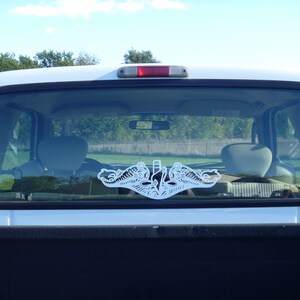 May include: A white truck with a white sticker of a submarine emblem on the rear window. The sticker is a silhouette of a submarine with two dolphins flanking the submarine.