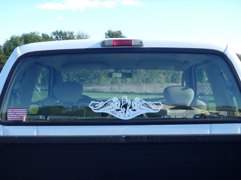 May include: A white truck with a white sticker of a submarine emblem on the rear window. The sticker is a silhouette of a submarine with two dolphins flanking the submarine.