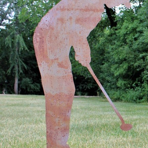 May include: A weathered metal sculpture of a golfer in a putting stance. The sculpture is a silhouette with a rusty brown finish, set on a grassy lawn with a backdrop of green trees. The golfer is bent over, holding a golf club.