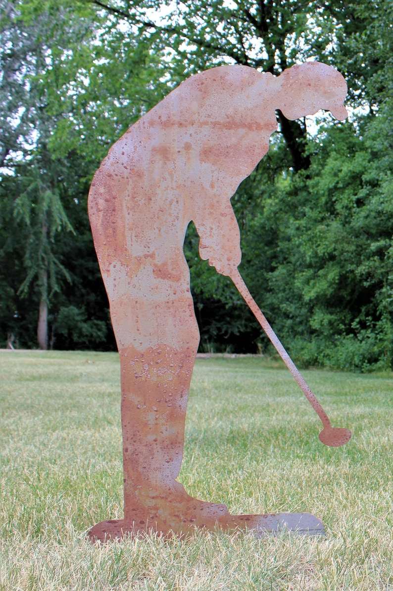 May include: A weathered metal sculpture of a golfer in a putting stance. The sculpture is a silhouette with a rusty brown finish, set on a grassy lawn with a backdrop of green trees. The golfer is bent over, holding a golf club.