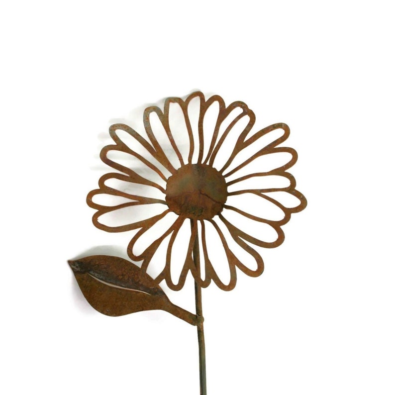 May include: A rustic metal daisy garden stake with a weathered, brown finish. The flower has a detailed, openwork design with a central disc and a single leaf. The stem is a simple, slender rod.