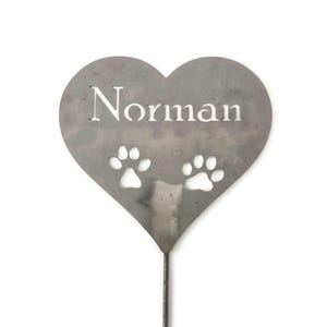 May include: A heart-shaped metal garden stake with the name "Norman" cut out in white. Two paw prints are cut out below the name. The stake is made of a silver-colored metal and is designed to be placed in a garden.