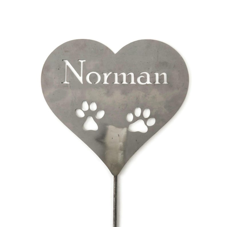 May include: A heart-shaped metal garden stake with the name "Norman" cut out in white. Two paw prints are cut out below the name. The stake is made of a silver-colored metal and is designed to be placed in a garden.
