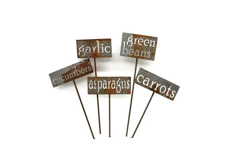 May include: A collection of weathered metal garden markers. Each marker displays a different vegetable name in white lettering: garlic, green beans, cucumbers, asparagus, and carrots. The markers are on thin metal stakes.