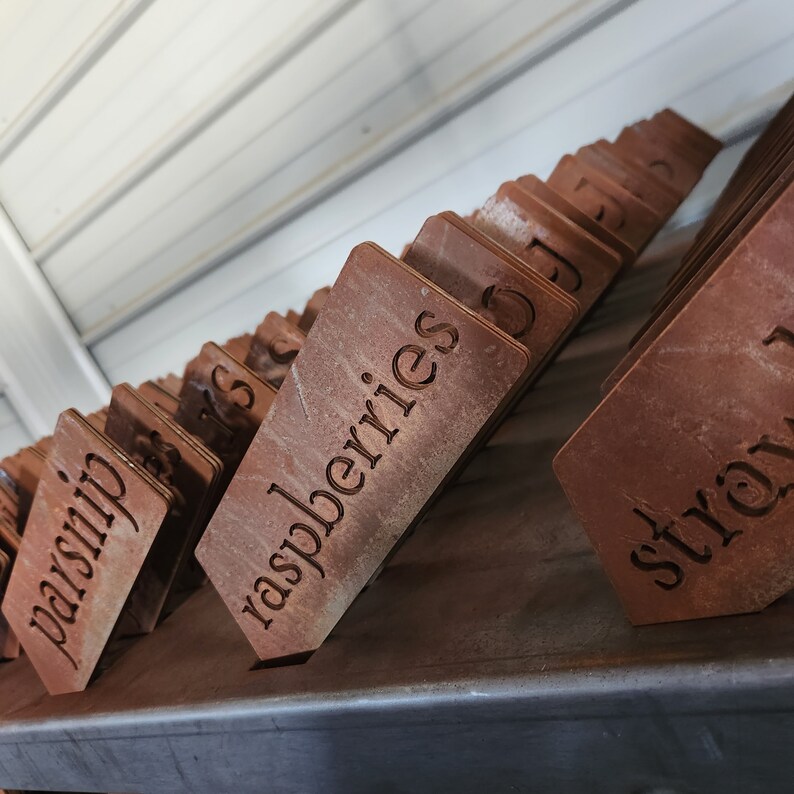 May include: Close-up of a stack of wooden plant markers with various labels, including "raspberries", "straw", and "parsnip". The markers are a rustic brown color and have a weathered look.