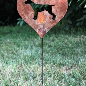 May include: A heart-shaped metal garden stake with a silhouette of a dog. The heart is made of rusty metal and has a dark silhouette of a dog in the center. The stake is in a grassy area.