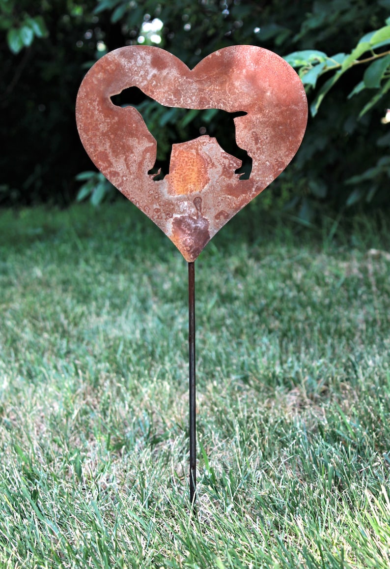 May include: A heart-shaped metal garden stake with a silhouette of a dog. The heart is made of rusty metal and has a dark silhouette of a dog in the center. The stake is in a grassy area.