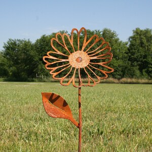 May include: A rustic, metal flower garden stake with a large, openwork daisy design. The flower and leaf are made of weathered, orange-brown metal, set against a backdrop of green grass and trees under a blue sky. A decorative outdoor accent.