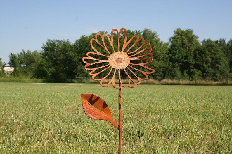 May include: A rustic, metal flower garden stake with a large, openwork daisy design. The flower and leaf are made of weathered, orange-brown metal, set against a backdrop of green grass and trees under a blue sky. A decorative outdoor accent.