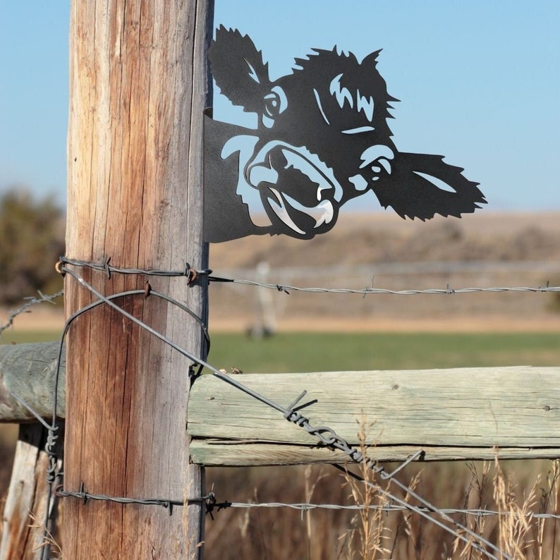 May include: A black metal silhouette of a cow's head peeking around a wooden fence post. The cow's head is facing the right side of the image and has a playful expression. The fence post is brown and weathered, with a wire fence running across the top.