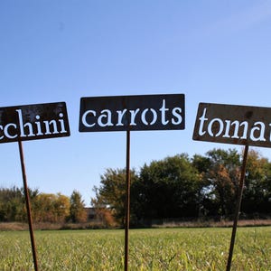 May include: Three weathered metal garden signs on stakes, each with white lettering against a dark background. The signs read "zucchini", "carrots", and "tomato", set in a grassy field under a blue sky.