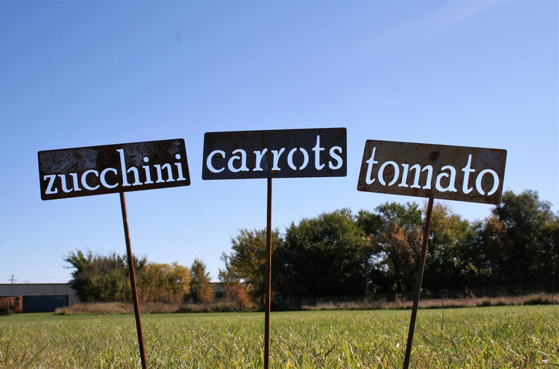 May include: Three weathered metal garden signs on stakes, each with white lettering against a dark background. The signs read "zucchini", "carrots", and "tomato", set in a grassy field under a blue sky.