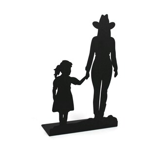 May include: A black metal silhouette sculpture depicting a tall figure wearing a cowboy hat standing next to a smaller child. The two figures are holding hands, and the entire piece is mounted on a flat, rectangular black metal base.
