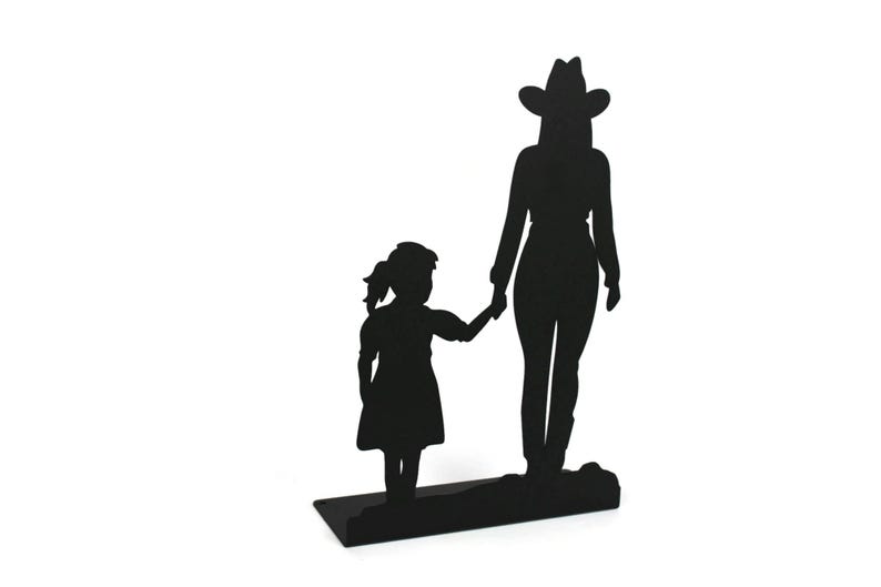 May include: A black metal silhouette sculpture depicting a tall figure wearing a cowboy hat standing next to a smaller child. The two figures are holding hands, and the entire piece is mounted on a flat, rectangular black metal base.