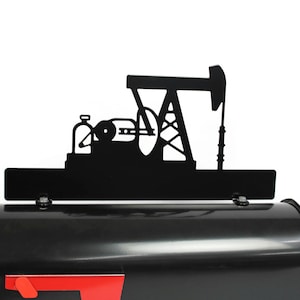 May include: Black metal mailbox topper depicting an oil pump jack silhouette. The topper is mounted on a black mailbox with a red flag. The design includes detailed cutouts of the pump jack's structure and machinery.