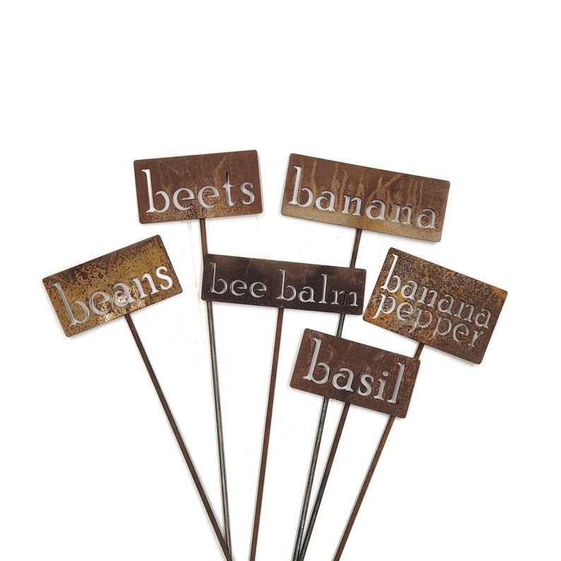 May include: A collection of rustic metal garden markers with the names of various plants, including "beets," "banana," "beans," "bee balm," "banana pepper," and "basil." The markers have a weathered, brown finish and are mounted on thin metal stakes.
