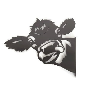 May include: Black metal wall decor featuring a cow's head with a playful expression. The cow's head is cut out of the metal, creating a silhouette effect.
