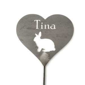 May include: A heart-shaped metal garden stake with the name "Tina" cut out at the top. A white silhouette of a rabbit is cut out below the name. The stake is gray and has a dark metal rod.