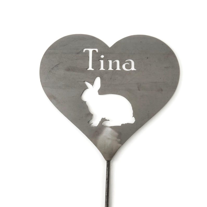 May include: A heart-shaped metal garden stake with the name "Tina" cut out at the top. A white silhouette of a rabbit is cut out below the name. The stake is gray and has a dark metal rod.
